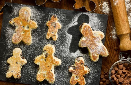 Aerial food photograph of homemade chocolate chip Welsh cakes baked in the shape of gingerbread men, dusted with icing sugar and served on a slate slab on a wooden table with cookie cutters, a rolling pin and chocolate chips