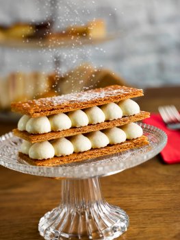 Food photograph close up of a mille feuille, laters of crisp golden puff pastry with vanilla creme patissiere sandwiched between them, dusted with icing sugar and served on a glass cake stand on a wooden table, with an afternoon tea stand in the background
