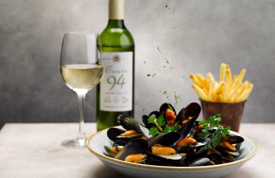 Food photograph of a bowl of steamed mussels in white wine sauce sprinkled with parsley, served on a grey background with a glass and bottle of white wine, and a portion of french fries in a copper pot