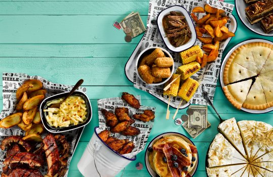 Food photograph of an Americana sharing table, a teal tabletop decorated with American flag pin badges and dollar bills, featuring shiny glazed barbecue pork ribs, potato wedges, macaroni cheese, barbecue chicken wings, bar marked grilled corn, pulled Southern style beef, fluffy pancakes with maple syrup, blueberries and bacon, key lime pie, apple pie, and chocolate and peanut butter layer cake