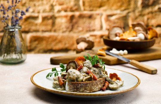 Food photograph of wild mushrooms in a cream sauce with parsley, spooned over a thick slice of golden sourdough toast, served in a home style setting with a bowl of wild mushrooms, and a small glass vase of lavender in the background