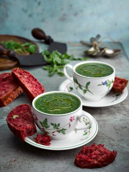 Food photograph of vintage teacups filled with homemade nettle soup and drizzled with olive oil, served alongside homemade beetroot and walnut bread and shot on a sky blue background with chopped nettles with a mezzaluna and a wooden chopping board