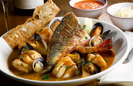 Food photograph of a traditional French bouillabaise, mussels, clams, prawns, cod chunks and a sea bass fillet in a tomato and wine sauce served with crisp golden toast shot in a restaurant setting