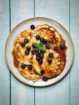 Food photograph of fluffy american style pancakes drizzled with maple syrup and topped with blueberries, served on a white plate on a blue wooden background