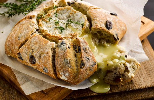 Food photograph of a baked camembert inside an olive pave, golden crusty bread studded with olives encasing a molten baked camembert which is spilling out where the bread has been torn, served on white wax paper in a rustic setting on a wooden table