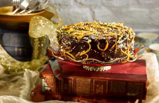 Food photograph of a homemade chocolate glazed cake, coated in shiny chocolate ganache and sprinkled with chopped pistachios and candied orange peel, served on a vintage glass cake stand in a vintage home style setting on a stack of books with gold fabric
