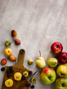 Aerial food photograph of autumnal fruits, large green bramley apples, red apples, bright green pears, various plums and greengages and scattered blackberries on a grey stone background