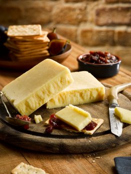 Food photograph of a block of crumbly cheddar cheese served in a rustic setting with chutney and crackers, along with a vintage cheese knife and teaspoon on a wooden board