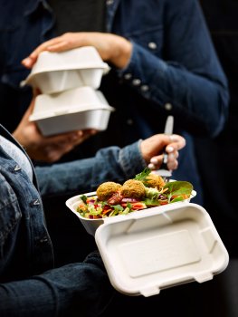 Food photograph of a crisp deep fried falafel salad in a compostable takeaway container with a forkful being lifted by a woman in a denim jacket, a man in a denim jacket holds two sealed containers in the background
