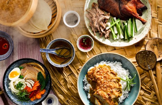 Food photograph of a Pan-Asian spread, a bowl of ramen noodles with roasted pork, soft boiled egg and vegetables, a breadcrumbed chicken katsu curry with steamed rice, and a crispy peking duck with spring onion, cucumber and pancakes in a steam basket, with pots of dipping sauce on a bamboo and pink wood background