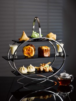 Food photograph of an Asian style afternoon tea, a three tier stand including steamed pork buns, chicken skewers, samosas, wontons, iced orange cake and lychee jelly pots, with an individual glass teapot shot in a dark hotel setting