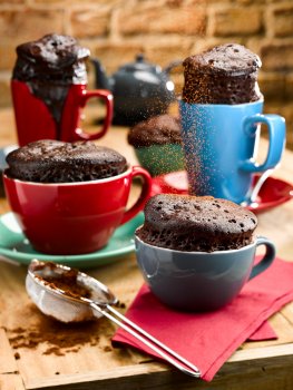 Food photograph of five chocolate cakes in coloured tea and coffee cups, on a wooden table with a brick wall in the background, with colourful saucers and napkins and cocoa powder being dusted over the top