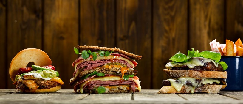 Food photograph of a trio of American sandwiches, featuring a pulled beef bagel with salsa, sour cream and guacamole, a Rueben sandwich with layers of pastrami and swiss cheese, and Russian dressing, and a vegetarian mushroom and cheese toast, with french fries. All of this on a bleached wooden tabletop with a dark wooden background