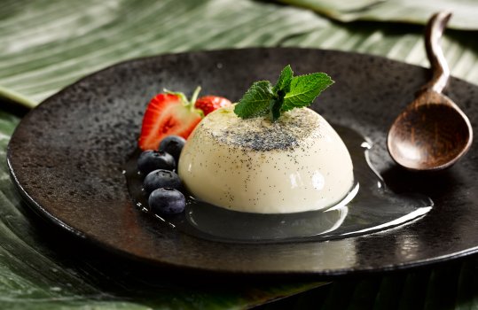 Food photograph of a vanilla and lime panna cotta, served on a black plate with fresh berries and syrup, shot in an Asian style setting with a carved wooden spoon on a banana leaf background