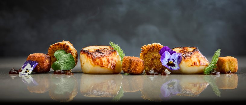 Food photograph close up of a fine dining starter, seared scallops served with crispy fried potato cubes and black pudding bonbons, decorated with edible flowers and served on a reflective silver background with clouds of smoke
