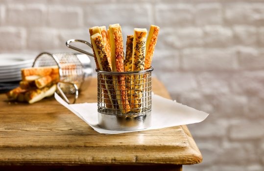 Food photograph of cheese and poppy seed breadsticks served in a steel basket on a sheet of wax paper, shot in a home style setting on a farmhouse table with white bricks in the background