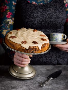 Food photograph of a homemade vegan pecan and maple cake, shot in a home style setting on a vintage cake stand being set down on a dark table by a woman in a floral dress