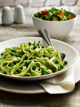 Food photograph of a pea and mint tagliatelle served in a white bowl in a restaurant style setting, with a white napkin and a bowl of mixed salad in the background, on a light grey wooden table with salt and pepper shakers