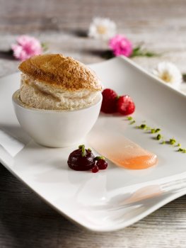 Food photograph of a fine dining dessert, a light fluffy peach souffle served with peach gel, raspberry syrup and chopped pistachios, on a light grey wooden background with flowers