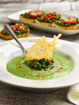 Food photograph of a pea and parmesan soup served in a white bowl with chorizo oil, fried peas and crispy chorizo and a parmesan crisp, with a platter of pea and tomato bruschetta in the background