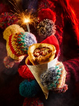 Food photograph close up of a pizza cone, a takeaway pizza with a cone shaped base filled with tomato sauce, cheese and pepperoni in a paper cone held by a girl wearing mittens and a woolen scarf holding a sparkler