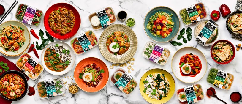 Food photograph of eleven plated ready meal dishes by City Kitchen, shot on a marble background alongside packaged ready meals and scattered ingredients such as cashews, pine nuts, paprika, kaffir lime leaves, chillies, coriander, sliced spring onion and red peppers