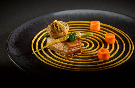 Food photograph of a fine dining main course, a slice of confit pork belly topped with roasted heritage carrot and a crispy black pudding bonbon wrapped in potato, served on a black plate with pickled carrots and a swirl of carrot puree, on a dark grey background