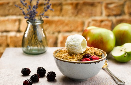 Food photograph of a golden crunchy apple and blackberry crumble served in a pale blue bowl and topped with a scoop of vanilla ice cream, the grey tabletop is decorated with scattered blackberries and large green apples in the background, along with a small glass vase of lavender