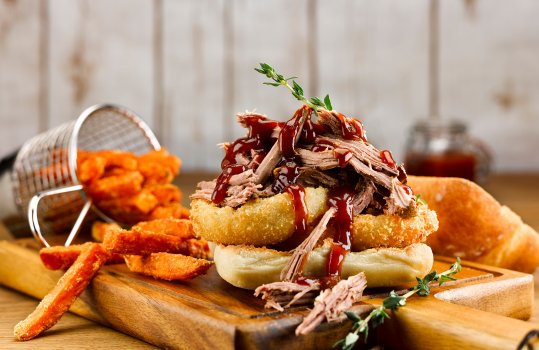 Food photograph close up of a Southern style pulled barbecue beef sandwich with crisp panko breaded onion rings and drizzled barbecue sauce served open faced on a wooden board with a basket of sweet potato fries