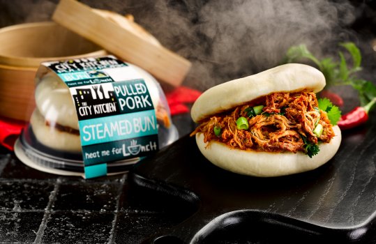 Food photograph of a ready meal pulled pork steamed bun, pulled pork in sweet and sour sauce with sliced spring onions and coriander in a fluffy steamed bao bun, served on shiny black tiles with a steam basket and billowing clouds of steam