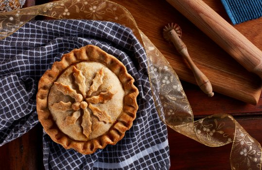 Aerial food photograph of a homemade festive raised pie, a crisp decorated pie shot from above in a home style setting, on dark wood with a rustic gingham tea towel and vintage baking tools including a rolling pin and pastry crimper