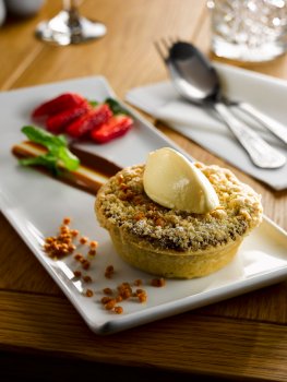 Food photograph of an individual apple pie with a crunchy crumble topping, served on a white plate alongside sliced strawberries and salted caramel pieces, topped with a quenelle of vanilla ice cream