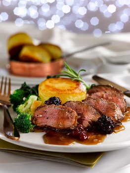 Food photograph of a juicy medium rare duck breast served on a white plate in a restaurant style setting with a fondant potato, steamed broccoli florets and blackberry sauce. Shot on a white background with glittering Christmas lights