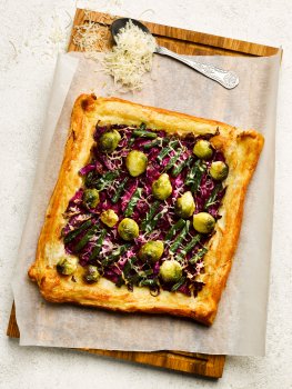 Aerial food photograph of a homemade Christmas puff pastry tart, a sheet of golden brown crispy baked puff pastry, topped with brussels sprouts, red cabbage, green beans and shaved parmesan, served on wax paper on a wooden board on a white tabletop, with a vintage spoon full of shaved parmesan