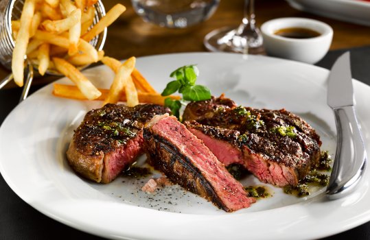 Food photograph of a juicy pink rare rib eye beef steak, served in a restaurant style setting on a white plate with a basket of french fries and chimichurri sauce, a glass of wine and a tomato salad are also on the dark wooden table in the background