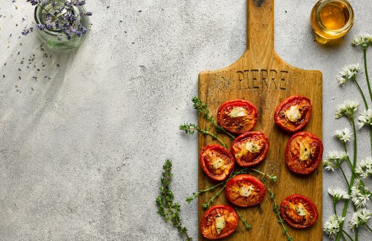 Aerial food photograph of roasted fresh tomatoes, topped with garlic and thyme and served on a wooden board alongside a glass jar of honey, wild garlic flowers and a small glass jar of lavender sprigs