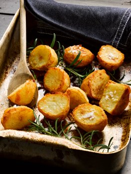 Food photograph of homemade crispy golden roast potatoes, shot in a home style setting in a rustic roasting tray with a vintage wooden spoon and tea towel, along with sprigs of rosemary, and topped with crystals of flakey sea salt