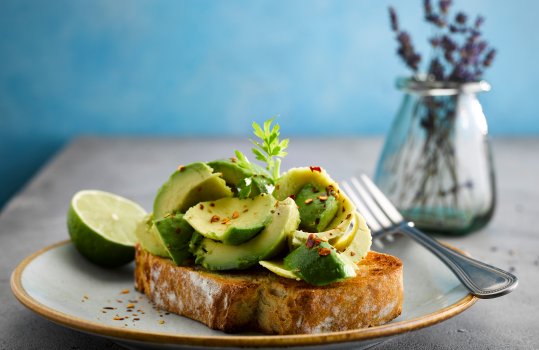 Food photograph of a slice of avocado toast, chunks and slices of avocado piled high onto a slice of golden toasted sourdough bread and sprinkled with chilli, served with a wedge of lime on a pale blue blate, on a grey tabletop with a small glass jar of lavender, and a bright blue background