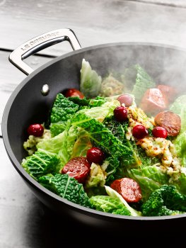 Food photograph close up of Christmas cabbage being cooked in a Le Creuset wok, fresh bright green savoy cabbage with fresh cranberries and chunks of crispy chorizo, topped with a mustard sauce shot in a home style setting on a grey wooden table