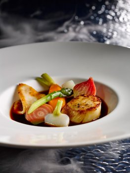 Food photograph of a fine dining starter, a fresh seared scallop served in a pool of consomme with steamed asparagus, carrot, beetroot and mixed radish, on a glass background with billowing clouds of smoke