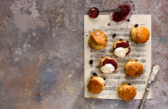 Aerial food photograph of homemade golden fruit scones, served with thick shiny homemade strawberry jam and rich clotted cream, served on a sheet of classical music on an abstract painted background