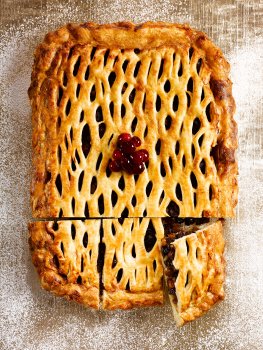 Aerial food photograph of a homemade sharing mince pie, crispy golden puff pastry encasing mincemeat filling and topped with a pastry lattice, topped with fresh redcurrants and icing sugar, three slices cut out and one is tilted upwards to show the filling