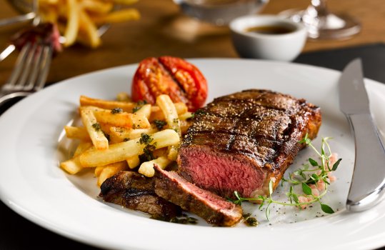 Food photograph of a juicy medium rare seared beef sirloin steak, served with french fries and a grilled tomato in a restaurant style setting - on a white plate garnished with herbs and pink Himalayan salt, on a wooden table with silverware and a wine glass