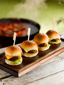Food photograph of four miniature slider burger canapes, mini brioche buns filled with miniature burger patties, lettuce, tomato and cheese, served on skewers, shot in an outdoor setting on a picnic table in front of a lit barbecue