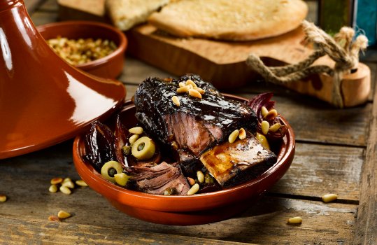 Food photograph of a braised beef short rib tagine, a shiny glazed beef rib in a ceramic tagine with pine nuts and olives, served with flatbread on a wooden board, on an antique wooden table with Moroccan lanterns