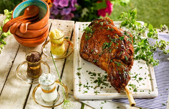 Food photograph of a roasted leg of Welsh lamb, served on a wooden board garnished with herbs, and served alongside various sauces in vintage teacups, shot in an outdoor setting on a picnic table with flowers and bushes in the background