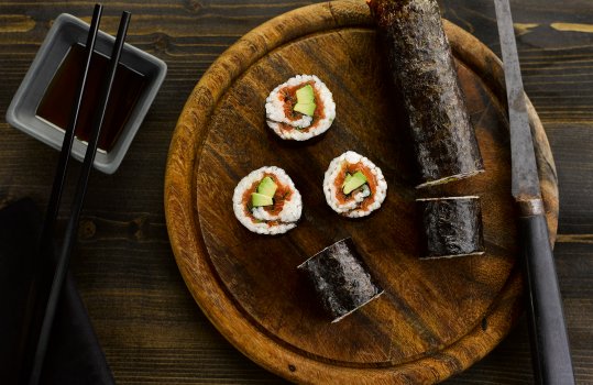 Aerial food photograph of a smoked salmon and avocado sushi roll, a whole sushi roll wrapped in nori shot on a wooden cutting board with a vintage slicing knife, with slices of the sushi roll stood on the board so show the salmon and fresh avocado filling. Shot on a dark wooden background with a dish of soy sauce and chopsticks