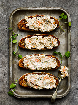 Aerial food photograph of smoked trout pate on toast, four slices of golden sourdough toast shot on a vintage baking tray, topped with flaked smoked trout in a cream cheese mix, garnished with watercress and shot on a grey stone background