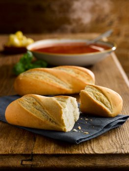 Food photograph of miniature baguettes shot on a vintage farmhouse table on a grey napkin, with a steaming bowl of tomato soup in the background along with herbs and curls of butter
