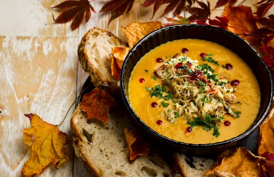 Aerial food photograph of a black bowl of bright orange butternut squash soup, topped with a crispy crouton, heaped with flaked white crab meat with chilli oil and herbs, shot on a weathered wooden background with slices of sourdough bread, and scattered autumn leaves